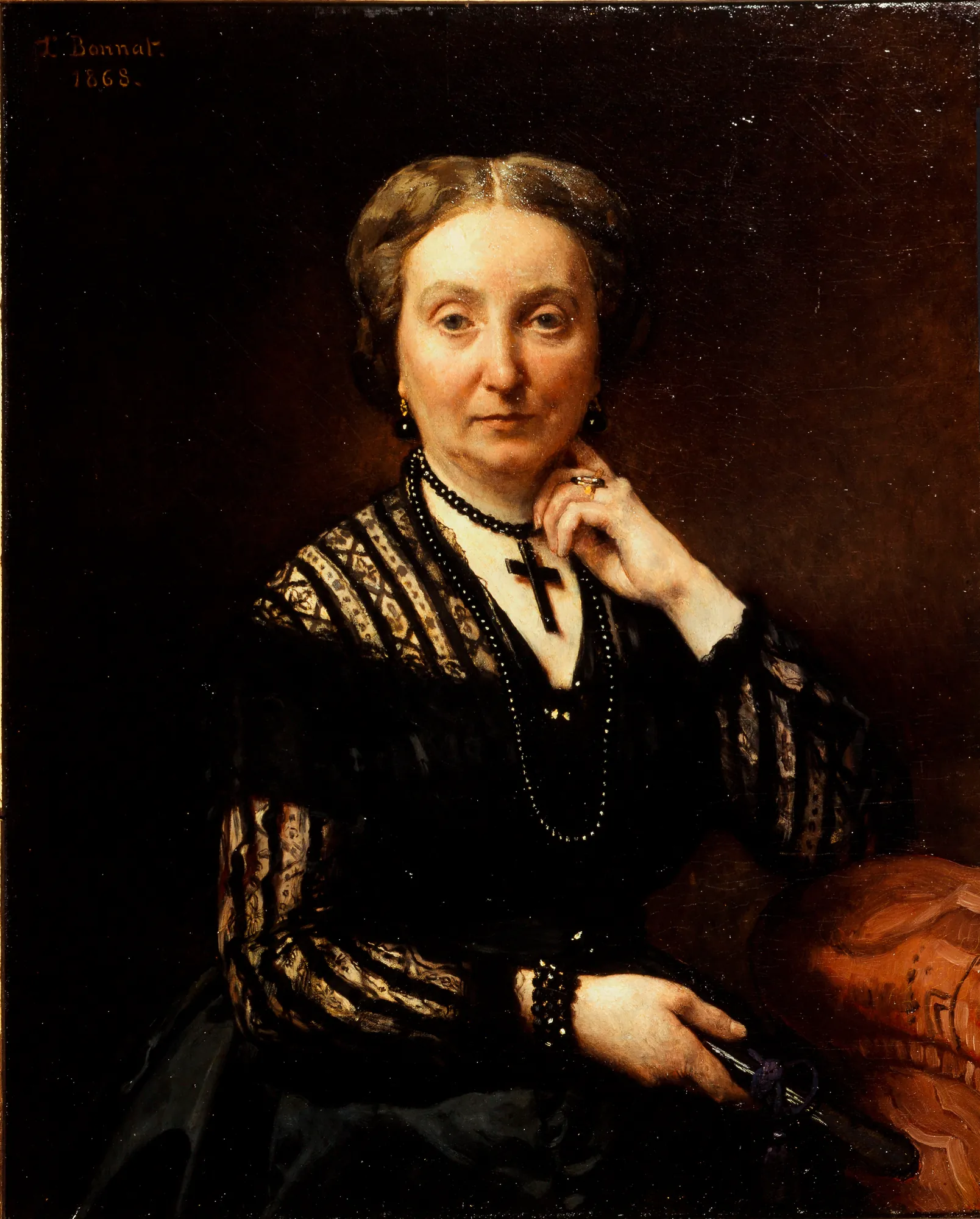 Portrait of a Woman Portrait of a Woman by Léon Bonnat. A waist-length portrait of a woman in a black lace dress, wearing a cross pendant and leaning against a red upholstered surface.