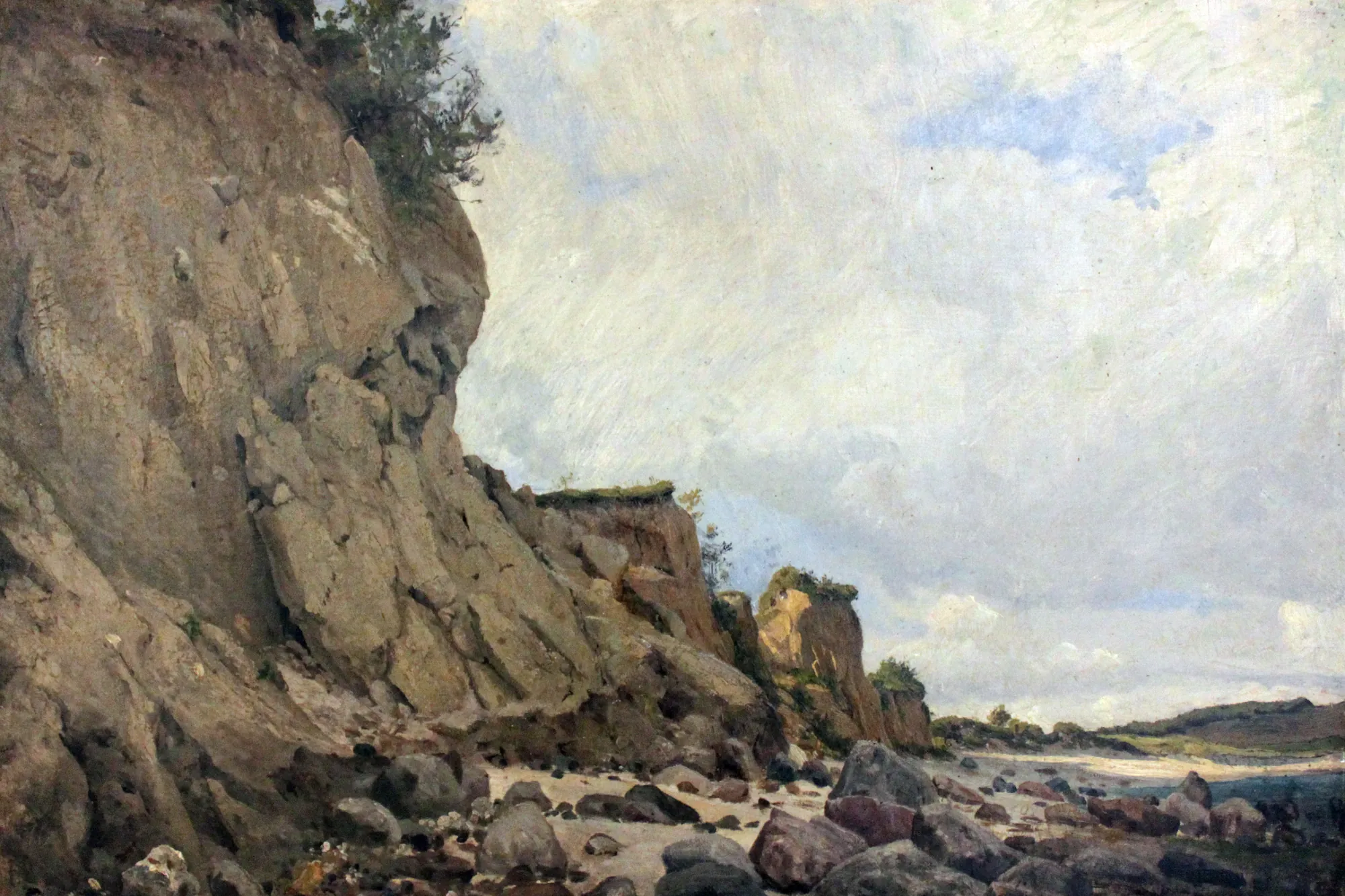 Beach near Mönchsgut Beach near Mönchsgut by Hans Peter Feddersen. A steep, rocky cliff looms over a boulder-strewn beach under a vast, cloud-filled sky.